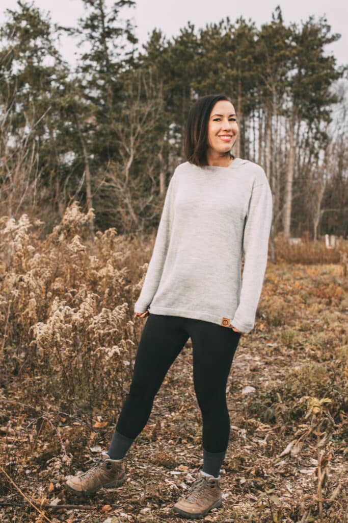 Lindsey wearing a grey alpaca hoodie, black leggings, and tan hiking boots in a wooded area during fall