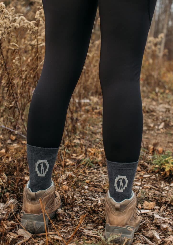 Wearing grey Paka socks that feature an alpaca on the back with black legging and tan hicking boots, standing on top of fallen leaves.