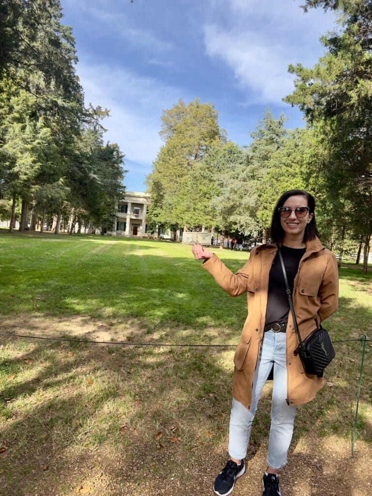 Lindsey wearing a tan jacket, black shirt and jeans holding up a $20 bill in front of Andrew Jackson's home