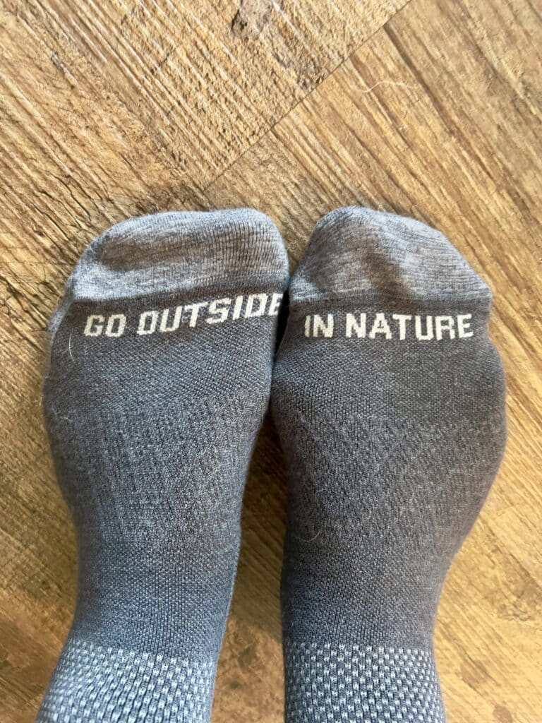 Grey socks that say "go outside" on sock and "in nature" on the other, set against hardwood flooring