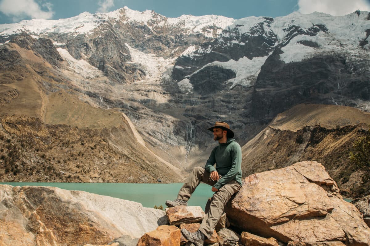Zac wearing an Oilskin hat, hiking shirt and hiking pants sitting on a rock beside Humantay Lake in Peru