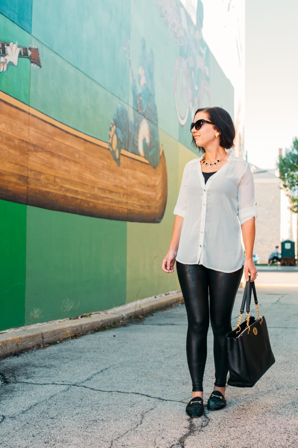 A woman with short dark hair wearing a white long sleeve blouse with faux leather leggings and black mules carrying a large leather black tote