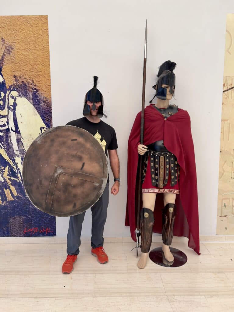 Zac wearing a Spartan helmet and holding a Spartan shield standing next to a Spartan mannequin in Greece 