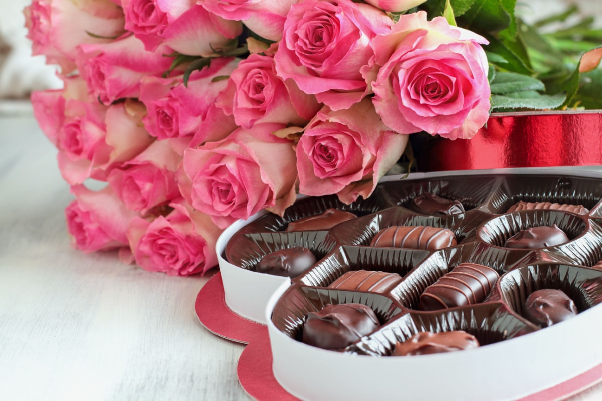 Dozen soft pink rose flowers with a heart shaped box of chocolate candy for Valentine Day over a wood background.