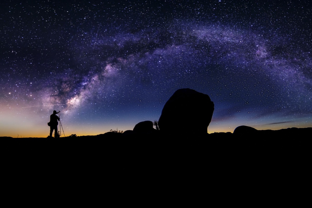 Photographer doing astro photography in a desert nightscape with milky way galaxy. The background is stary celestial bodies in astronomy. The heaven depicts science and the divine.