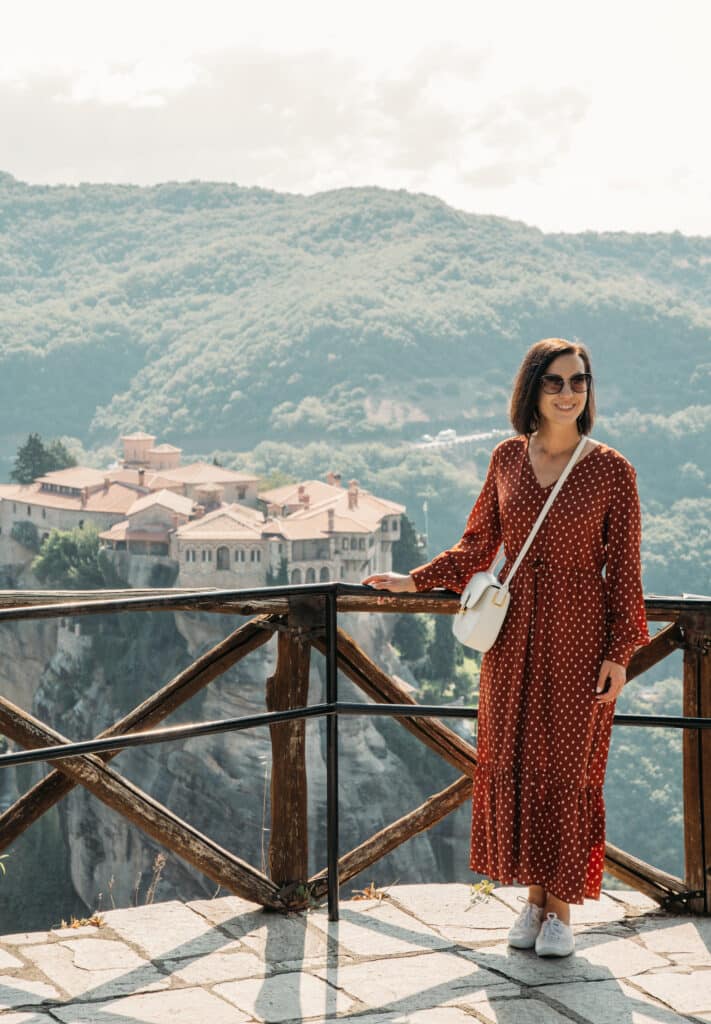 Lindsey of Have Clothes, Will Travel wearing a maxi dress and white Keds Sneakers with a white designer bag. She is standing by a railing with a monastery in the background at Meteora Greece