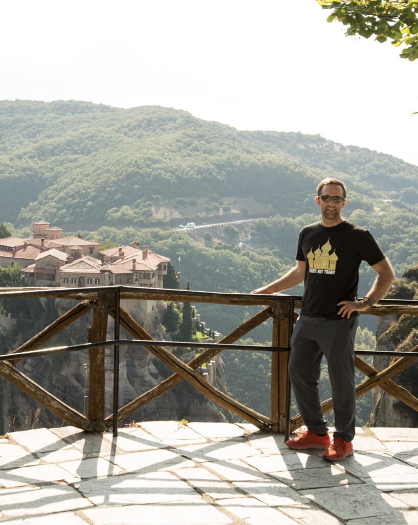 Zac of Have Clothes, Will Travel is wearing a black t shirt, hiking pants, orange sneakers and is standing by a railing with a monastery in the background at Meteora Greece