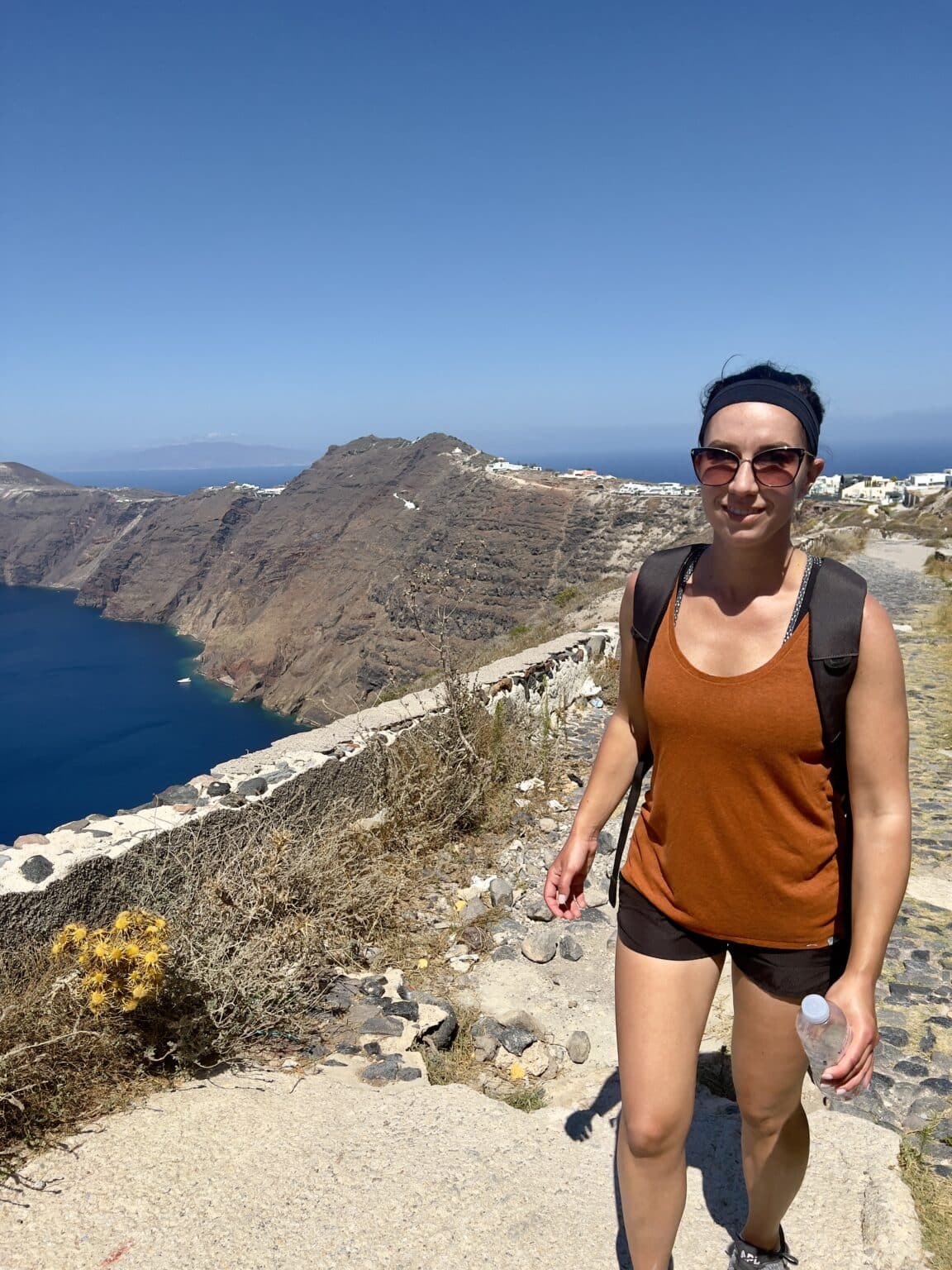 Lindsey wearing a burnt orange tank top and black shorts hiking in Santorini on a clear sunny day