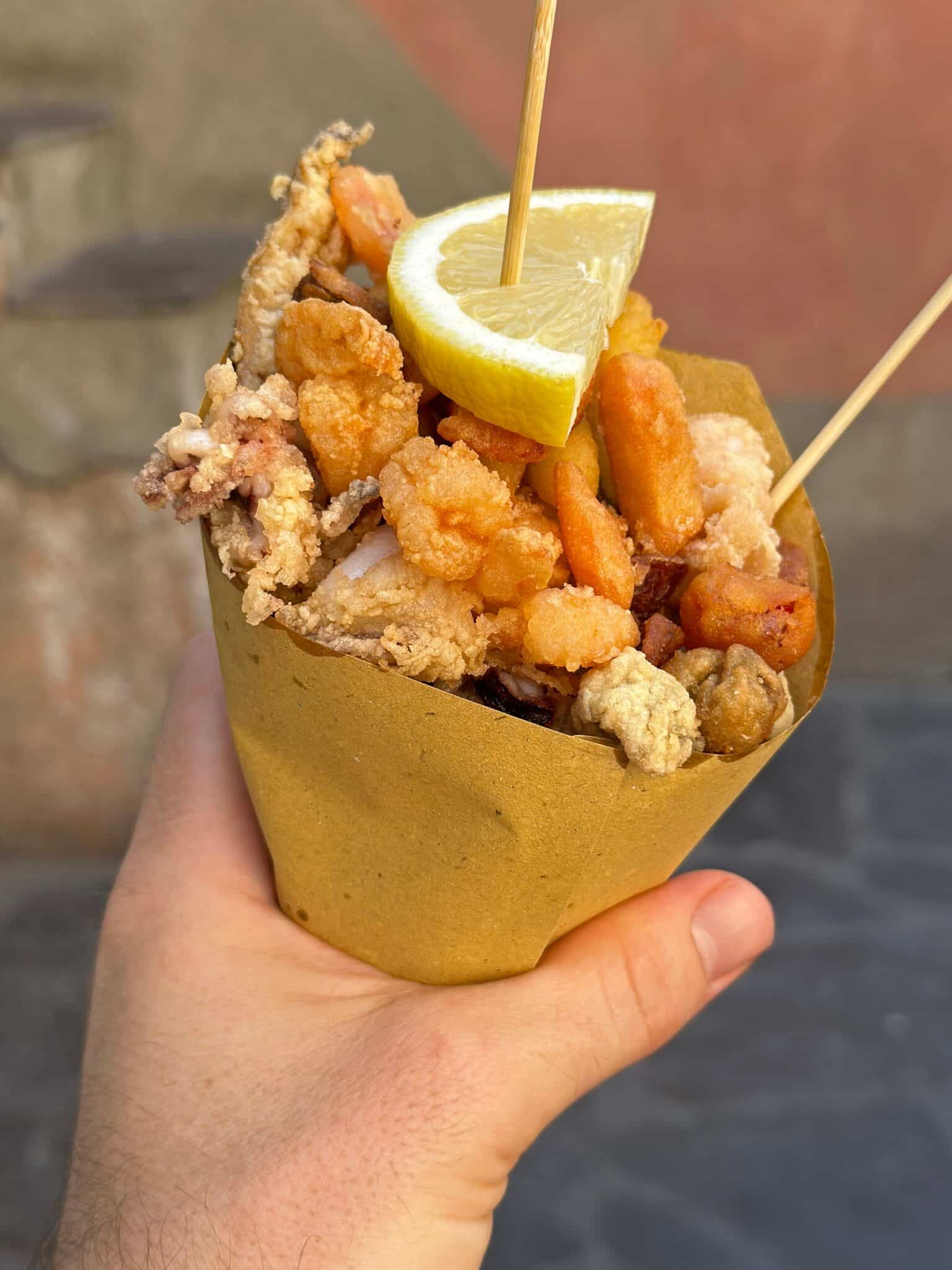 A hand holds a cone of fritto misto, which is an Italian street food. This particular version includes fried calamari, anchovies, shrimp, and vegetables, topped with a lemon to squeeze.