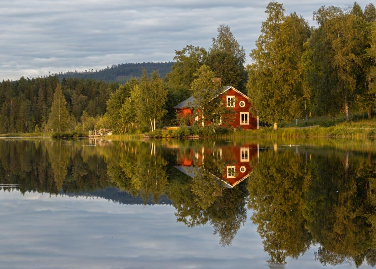 Swedish stuga by a calm lake on a cloudy summer day. The sunlight illuminates the green trees and the red house, while the interesting cloud-filled sky covers the heavens. All reflected in the water