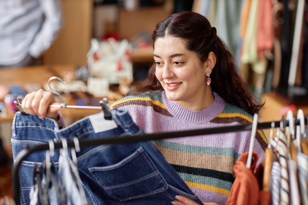 Portrait of smiling Middle Eastern female customer holding hanger with second hand pants choosing vintage gently used clothes at thrift store, copy space