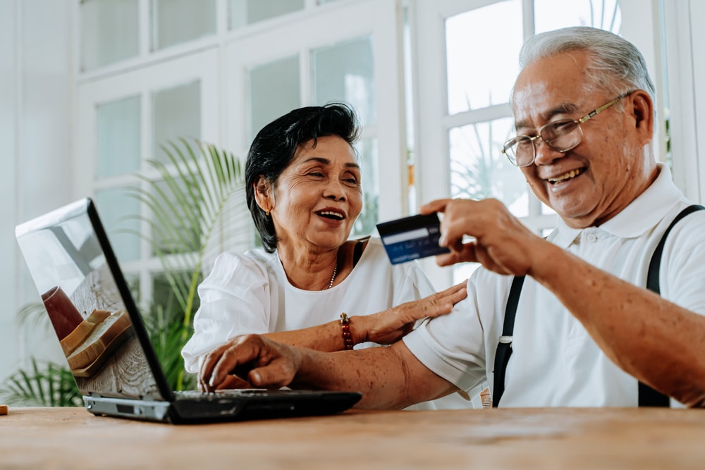 Delighted Asian man and woman using laptop and credit card to make order in online shop while sitting at table at home. Senior couple doing online shopping