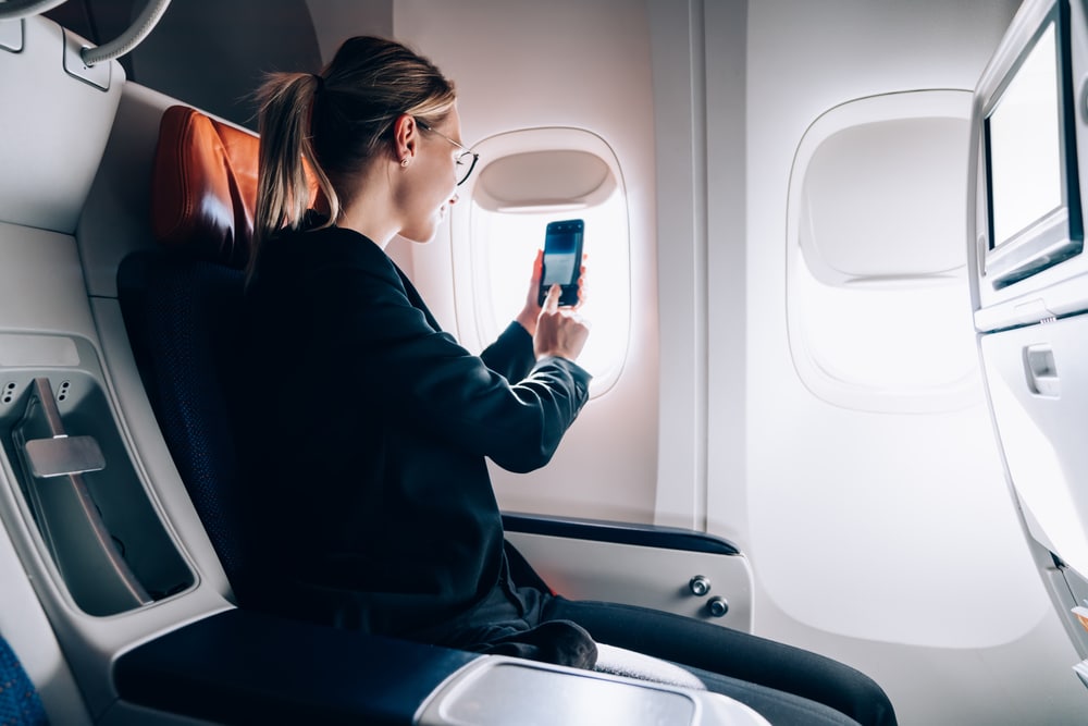 Millennial passenger using smartphone camera for shooting video vlog from window porthole in jetliner airplane during international comfortable flight in business class, woman photographing view