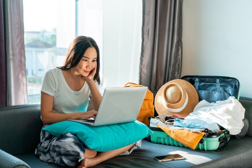 Asian woman prepare things into baggage for vacation trip at home. Happy female using computer to planning travel trip and booking tickets or hotel room on web. Travel, summer and holiday concept.