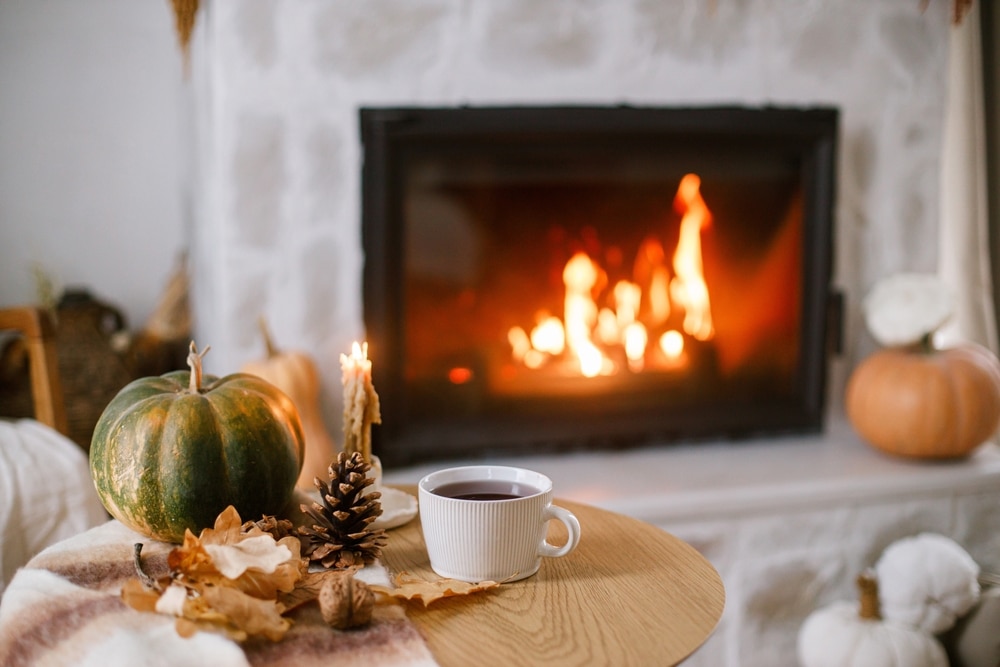 Warm cup of tea, pumpkin, autumn leaves on wooden table on background of burning fireplace. Cozy autumn still life. Thanksgiving and Halloween. Fall hygge