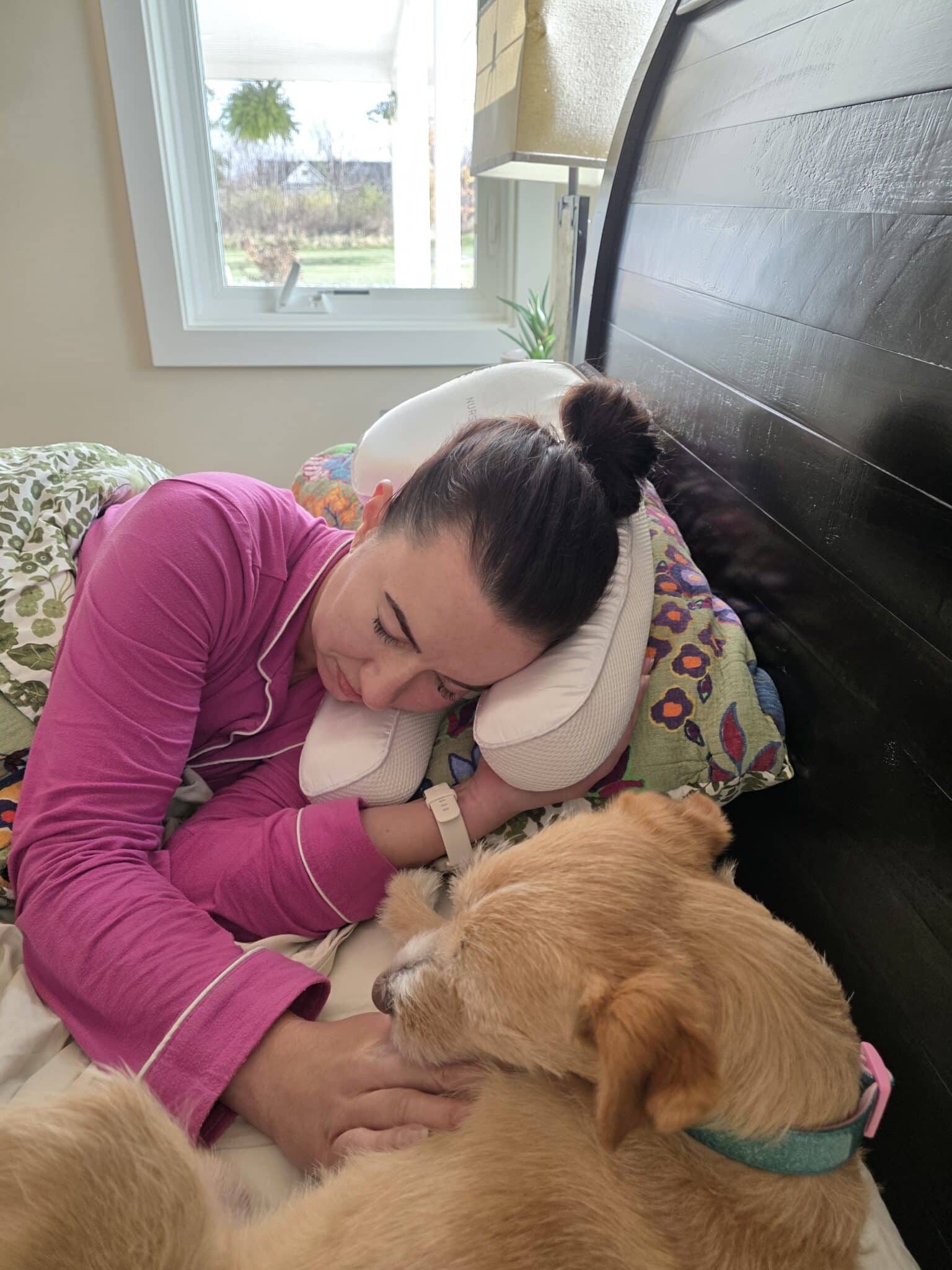 Lindsey using her Nurse Jamie Beauty Bear Memory Foam Pillow wearing pink pjs, her small dog sleeps next to her