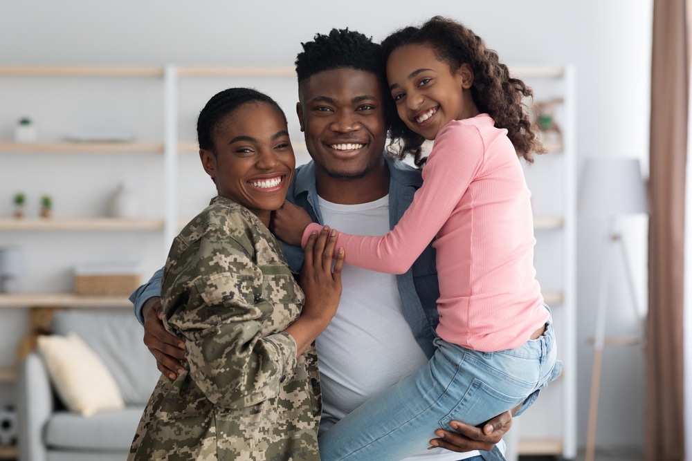 Happy african american woman in military uniform posing with her family at home, female soldier reunited with her husband and daughter, embracing all together and smiling at camera, copy space