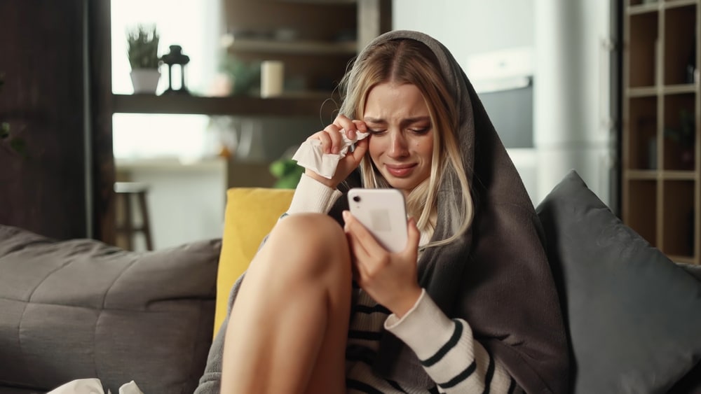 Portrait of crying young blond woman with smartphone sitting on the couch under the blanket and reading bad news email break up message from ex-boyfriend or personal problems at home