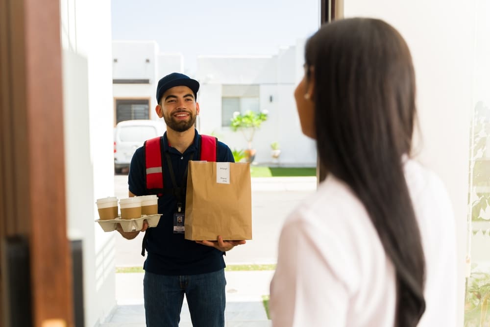 Happy delivery man holding a paper bag with takeout food and coffee for a client at the front door of her house