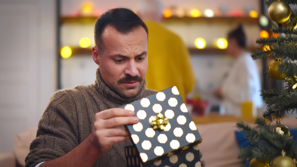 Portrait of handsome man opening christmas present and feeling disappointed. Guy standing near christmas tree unpacking gift and looking unhappy