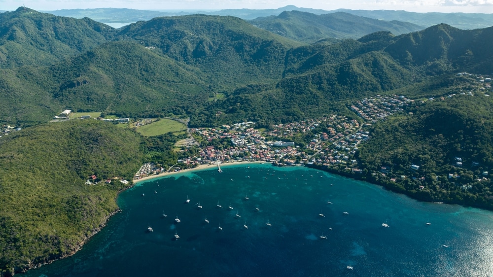 Drone picture of Anse d'arlet, its beach, church and jetty in Martinique, Caribbean island, with boats, houses, turquoise water and a cloudy blue sky in the background.