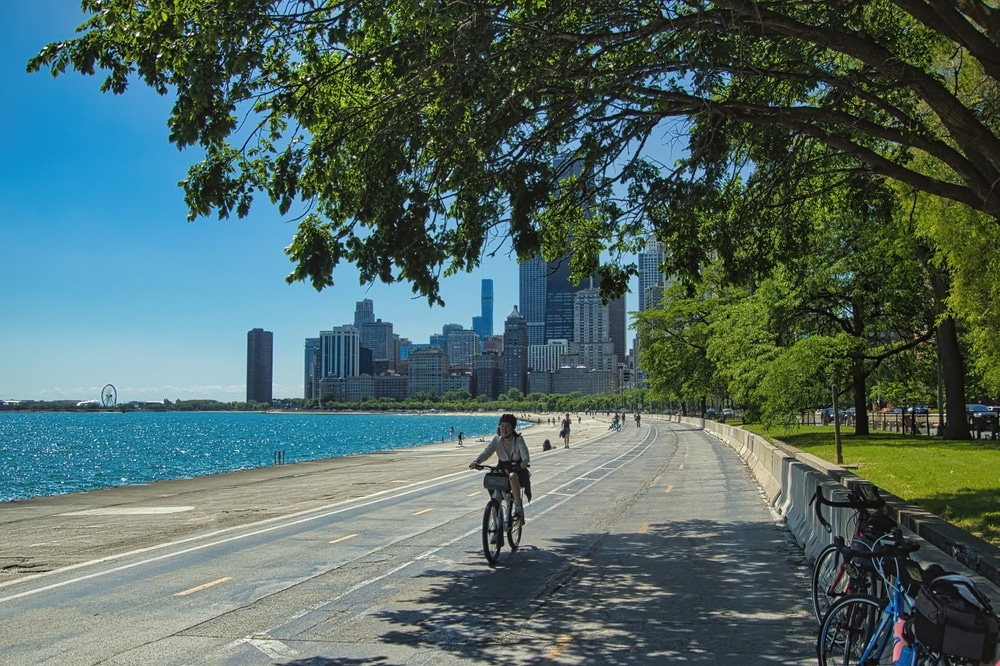 Chicago, Illinois, USA - May 28, 2024: Sunny early Summer landscape of a cyclist riding beside Lake Michigan on the Chicago Lakefront Trail with tall buildings in the background.