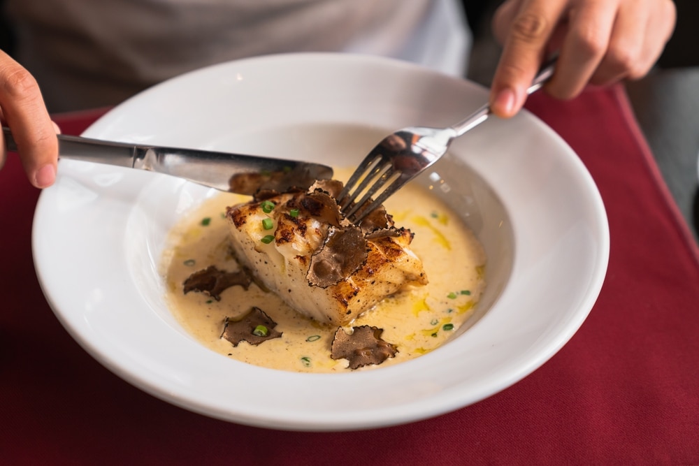 Hands of young man with knife and fork cutting fish steak and cream sauce garnished with slices of truffle on white plate while having lunch by served table