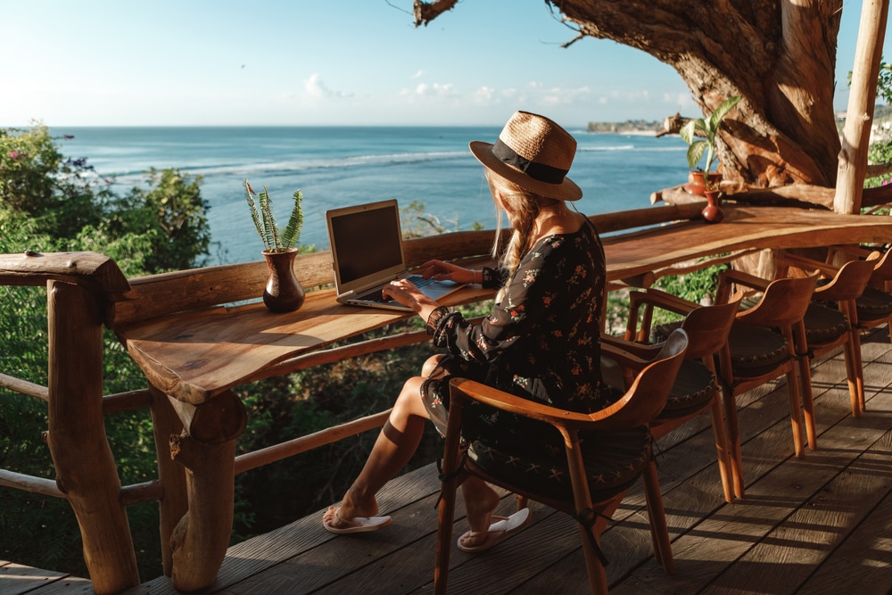 Freelance concept. Pretty young woman using laptop in cafe on tropical beach in outdoor cafe terrace with sea view. Work and travel