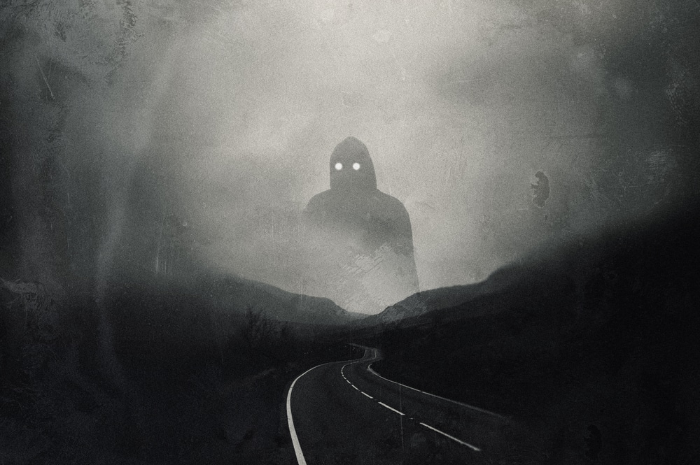A grainy horror concept, of a giant hooded figure with glowing eyes standing above a mountain road. With an abstract blurred edit.