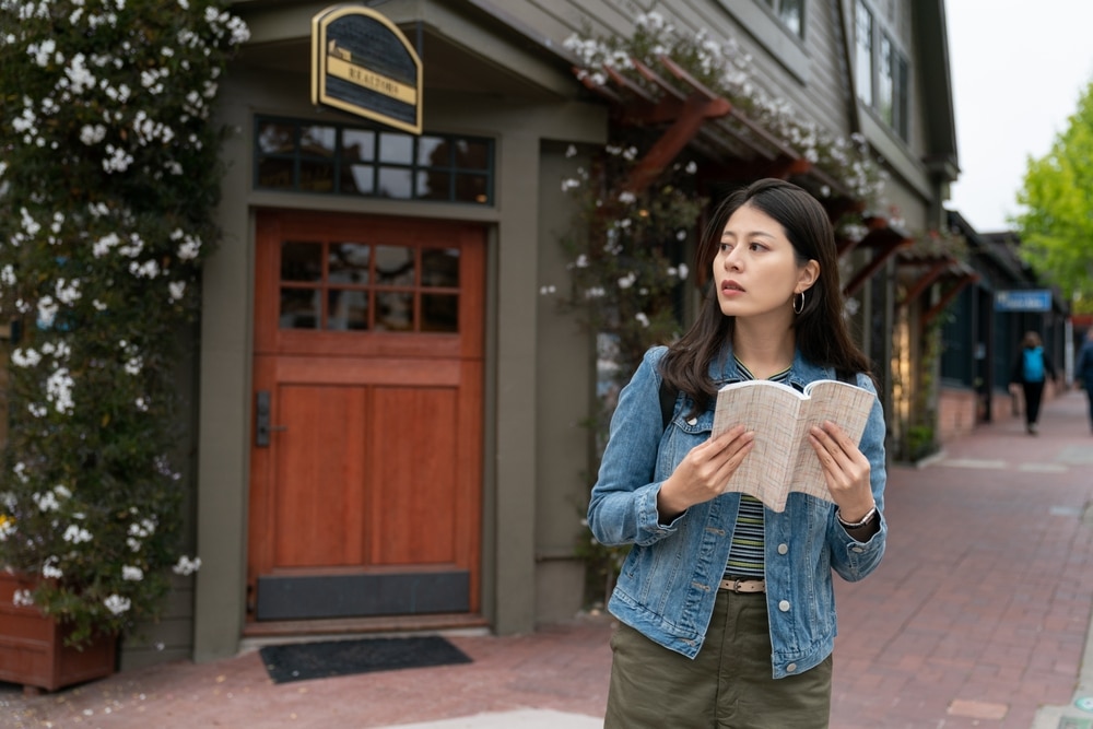 asian woman visitor holding guidebook and looking away into space on the red brick sidewalk near the entrance door of a wooden boutique shop in Carmel by the sea California usa