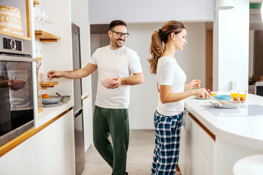 A woman in pajamas is preparing breakfast while a man is looking at her and helping her.