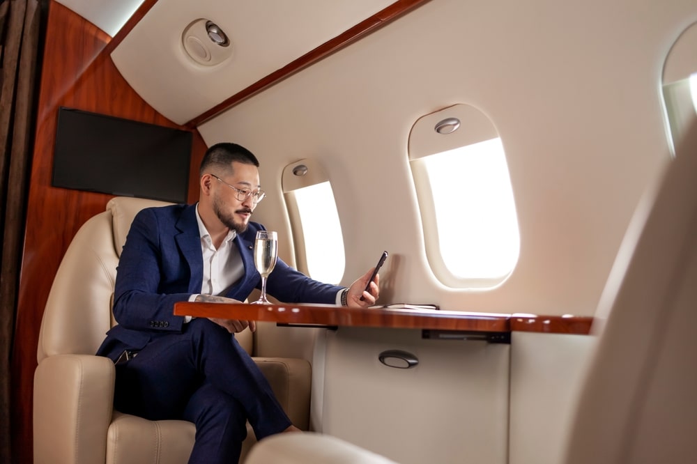 asian businessman in suit and glasses with glass of champagne flies in private luxury jet and uses smartphone, korean entrepreneur resting and looking at the phone in flight, luxury lifestyle