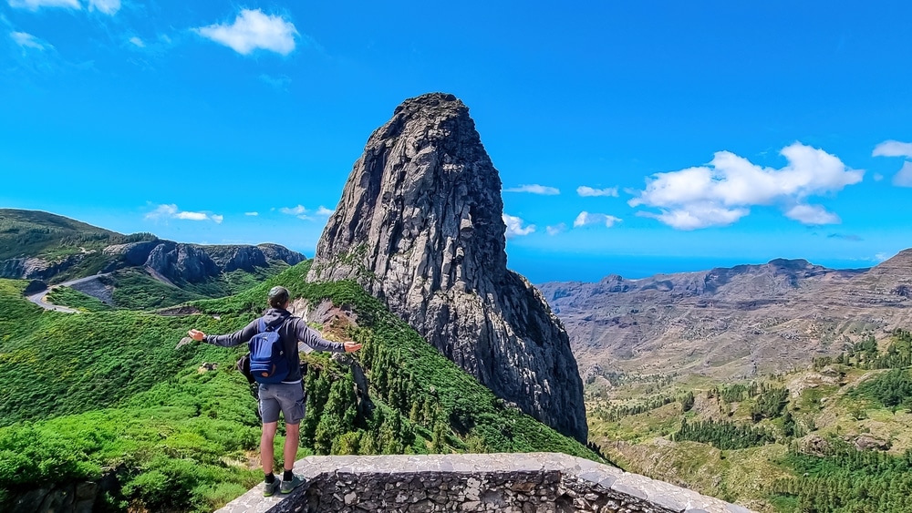 Sport man with backpack standing in front of massive volcanic rock formation Roque de Agando in Garajonay National Park on La Gomera, Canary Islands, Spain, Europe. Hiking trail on sunny day in summer