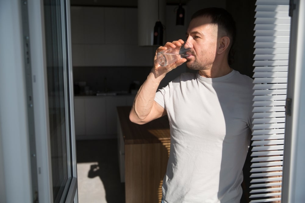 A 40-year-old man in a white T-shirt drinks water of their glass in the morning. Healthy lifestyle
