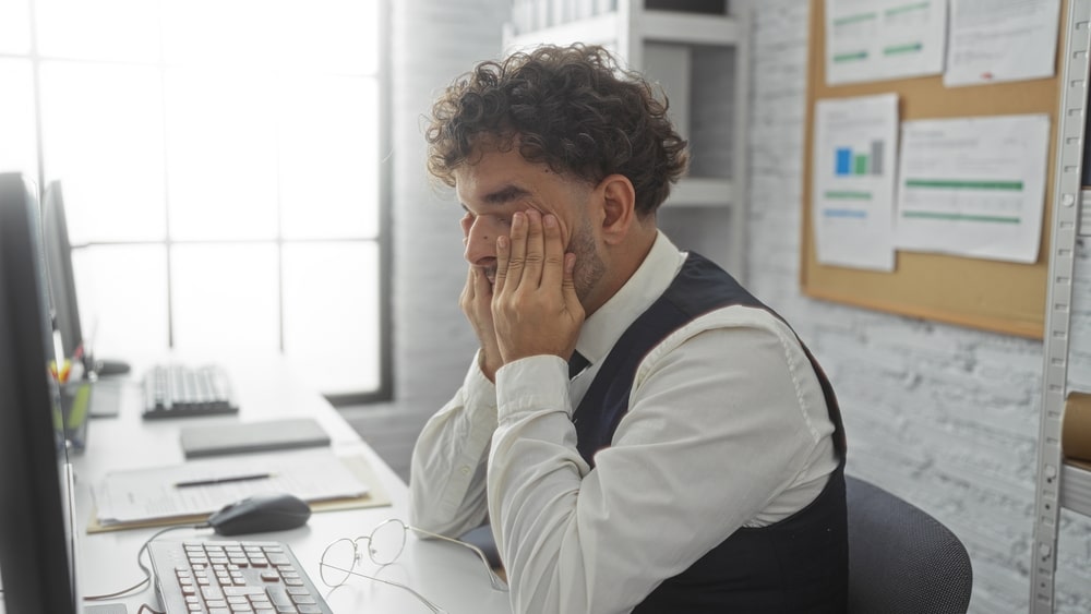Tired hispanic man in office rubbing eyes sitting at desk wearing vest working indoors beside computer in modern workplace with documents on pinboard