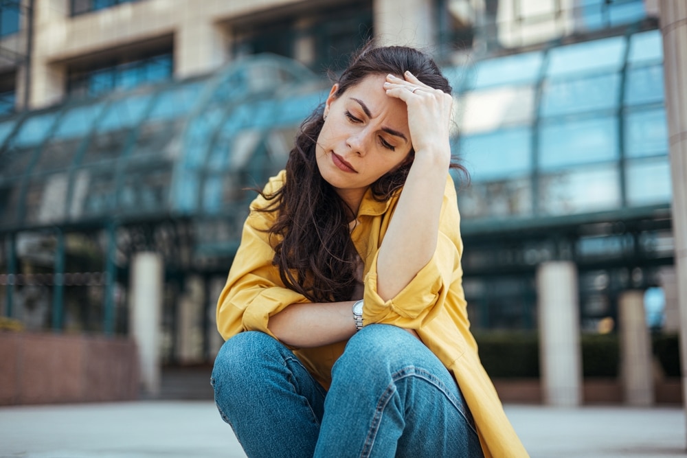 A contemplative young woman sits in a cityscape setting, conveying emotions of thoughtfulness and introspection amidst a modern architectural background.