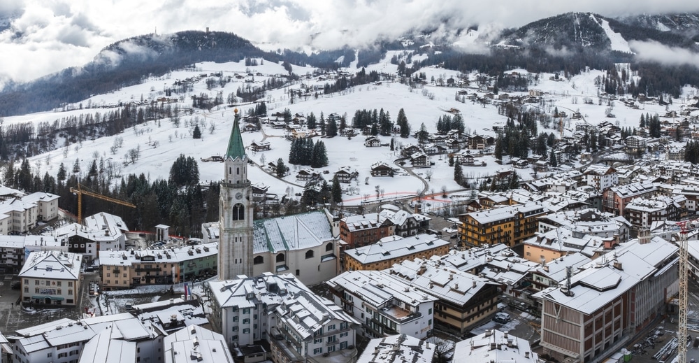 Aerial view of Cortina d'Ampezzo, Italy, featuring the Basilica Minore dei Santi Filippo e Giacomo, snow covered rooftops, and misty Dolomite peaks.