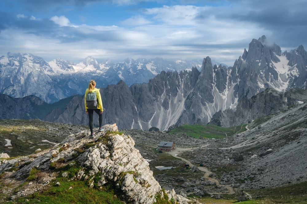 Young woman hiker with backpack standing on a rock in majestic mountain valley with high alpine mountain peaks, cloudy sky at sunset in summer. Hiking in Dolomites, Italy. Trekking, travel in spring