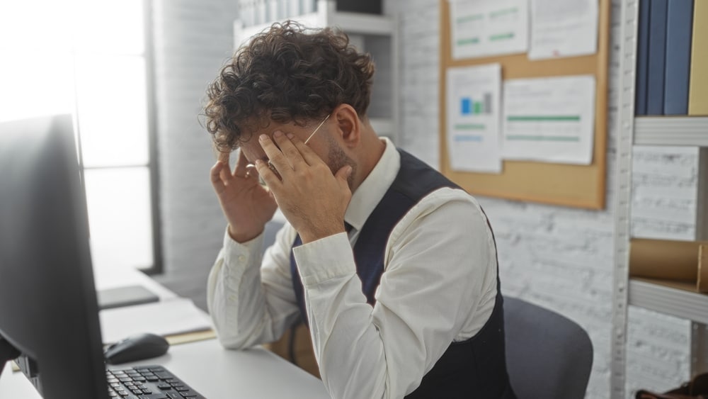 Frustrated young man in formal wear sitting at desk in modern office interior with hands on face indicating stress amidst paperwork and computer