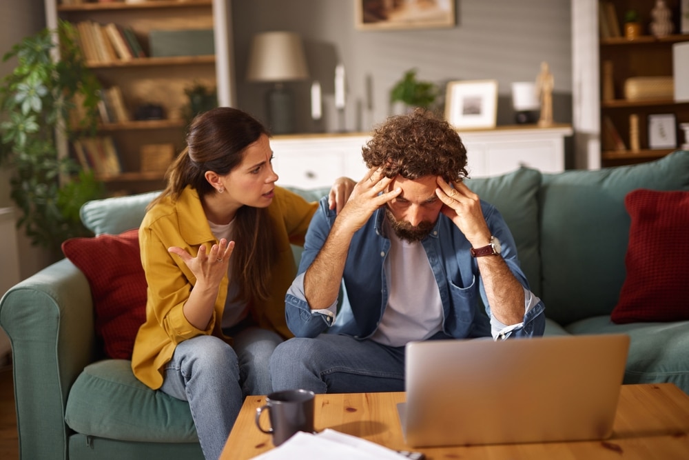 A woman expresses concern to a man sitting on a couch, who appears stressed while looking at a laptop. Papers and a coffee cup are on the table, highlighting their financial struggle.