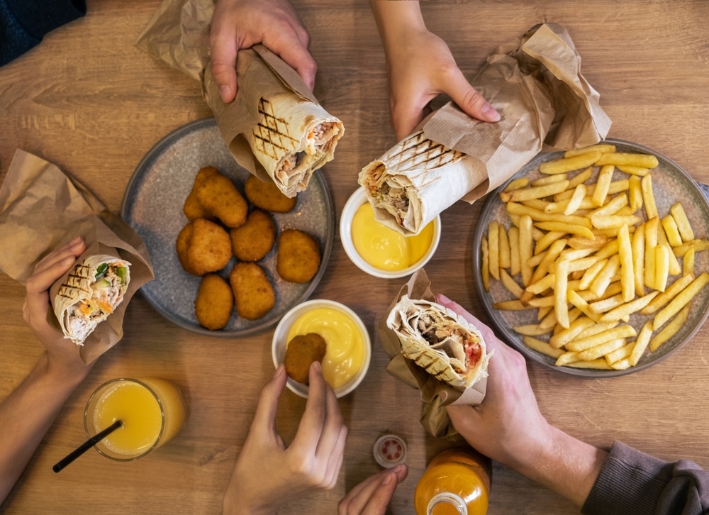 Hands of various individuals are enjoying flavorful wraps, crispy fries, and golden nuggets on a wooden table, showcasing a lively and appetizing meal setting