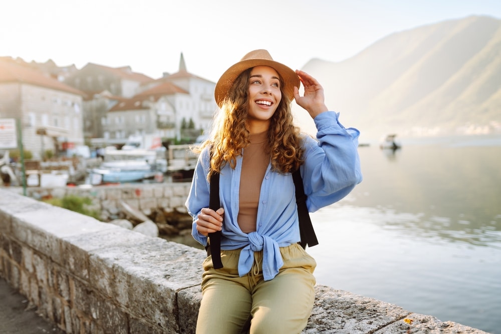 A cheerful woman poses by the water in a charming coastal town, wearing a hat and a light blue blouse, surrounded by serene mountains and boats. Europe travel.