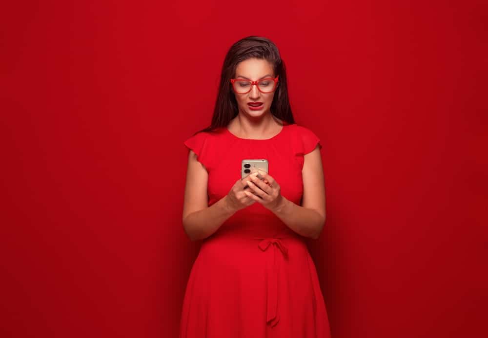 Impatient young woman with the mobile phone in the trouble, no time concept, isolated on red background. Worried girl looking to phone. Studio shot
