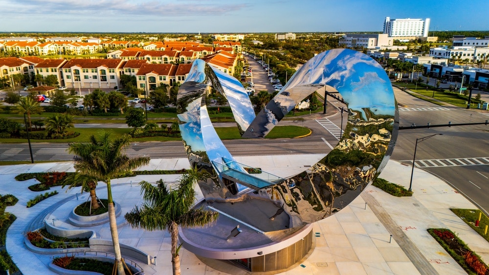 Port St. Lucie, Florida, United States – October 10, 2025:
Aerial view of a polished stainless steel heart-shaped public art sculpture at the entrance of the Traditions master-planned community in Port St. Lucie, Florida. The reflective metal surface mirrors surrounding palm trees, roadways, and nearby residential and commercial buildings.