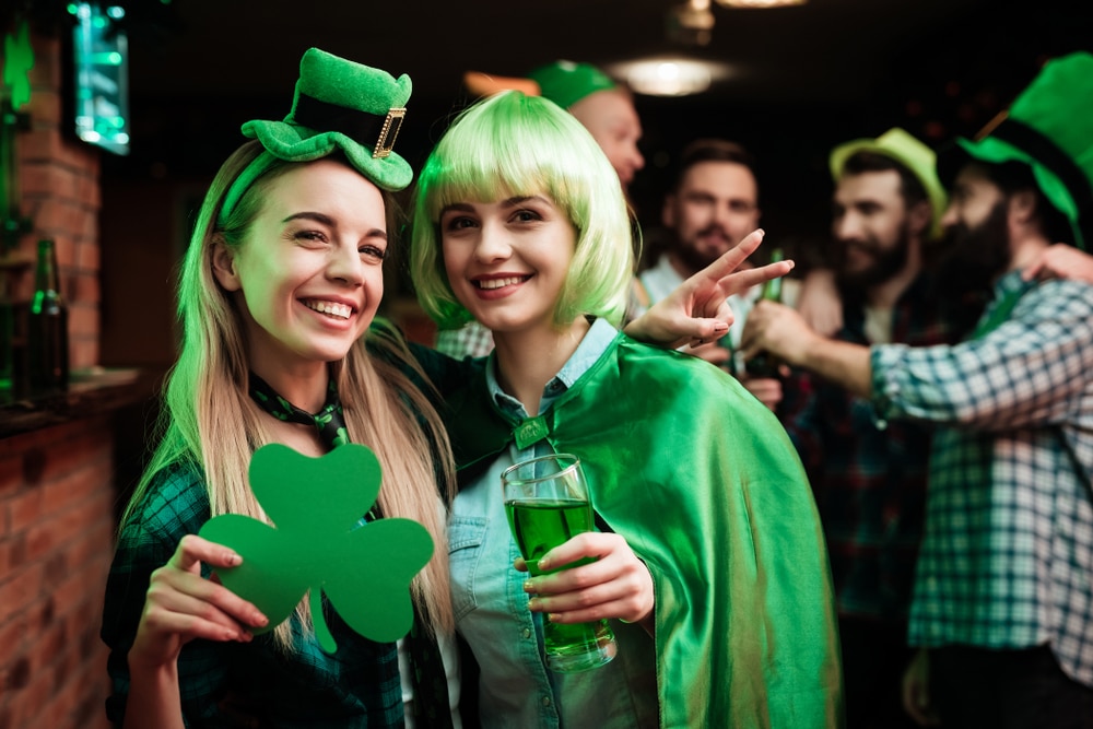 Two girls in a wig and a cap are photographed in a bar. They celebrate St. Patrick's Day. They are having fun. One girl is holding a clover.