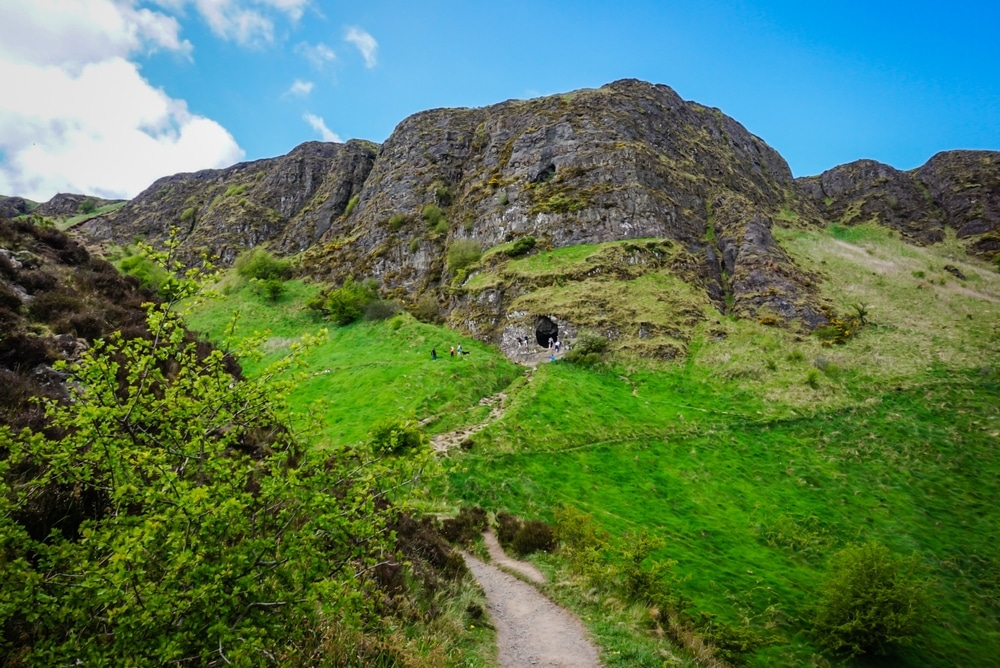 A stunning view of Cave Hill near Belfast, Northern Ireland, featuring a hiking trail winding through vibrant green grass towards a natural cave entrance on the rugged hillside. This scenic landscape