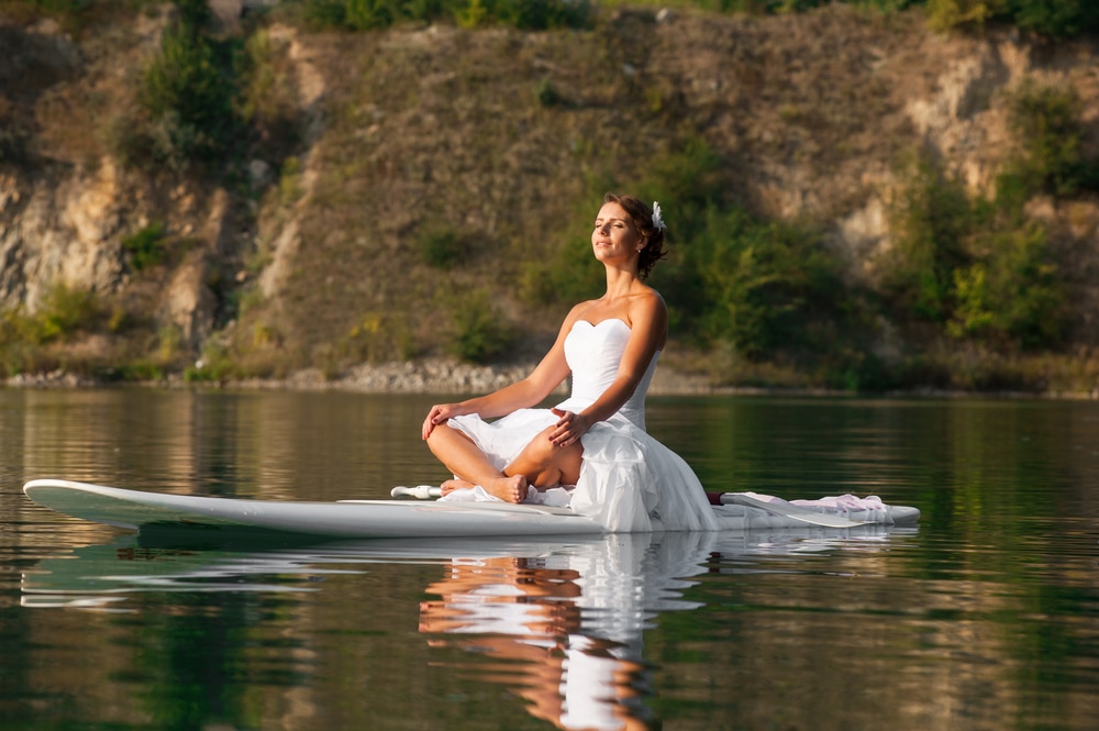 Girl in white dress meditating, sitting on a paddle board on water