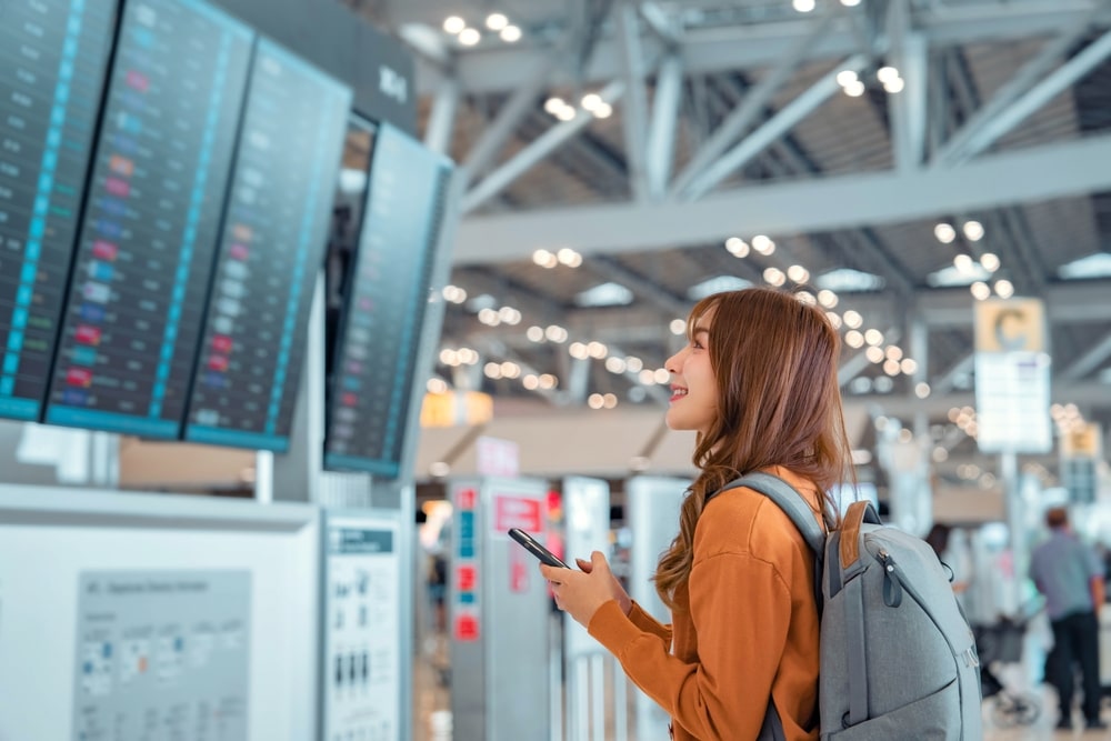 Happy asian woman traveller checking flight schedule departures board in airport terminal hall in front of check in counters. Tourist journey trip concept.