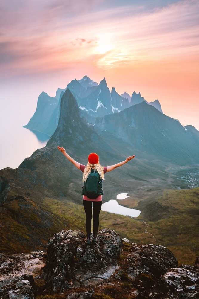 Traveler woman with backpack hiking in Norway girl raised arms alone on mountain top outdoor travel summer vacations active healthy lifestyle success motivation concept explore Senja island