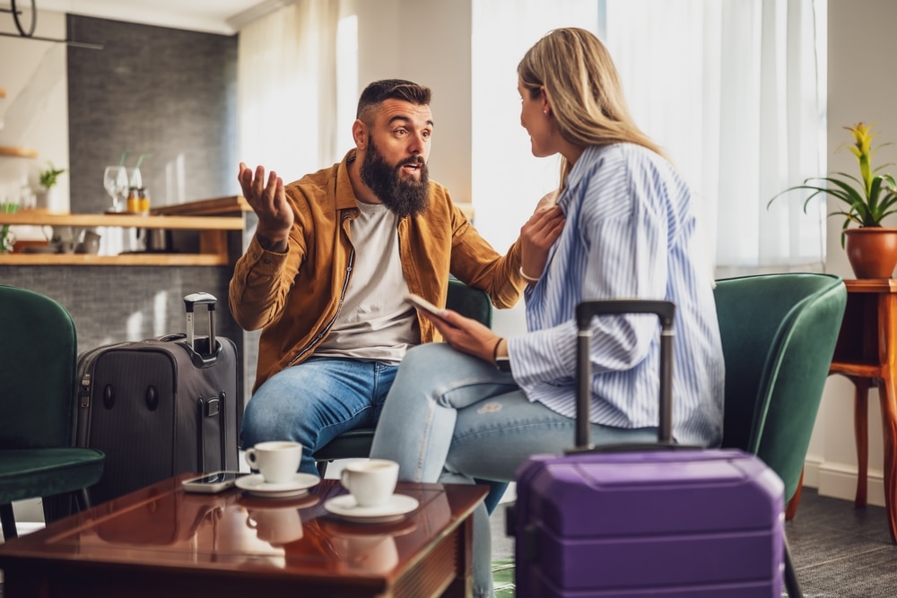 Couple is sitting at airport cafe before check in. They are going on vacation. Man is worried and frustrated.